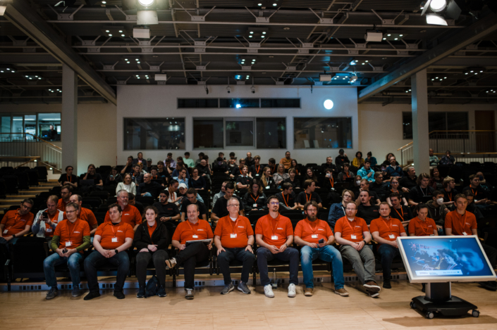 PIT Hackathin-Publikum mit Teilnehmenden in orangenen Shirts in einer IT-Eventhalle